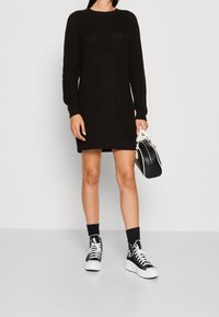 Robe noire en tricot à manches longues et col rond, associée à des baskets blanches et à un petit sac noir et blanc.