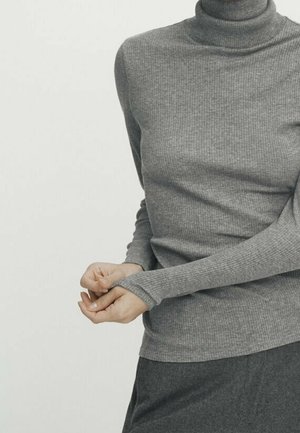 Personne portant un pull à col roulé côtelé gris ajusté et un pantalon gris foncé, ajustant la manche avec ses mains.