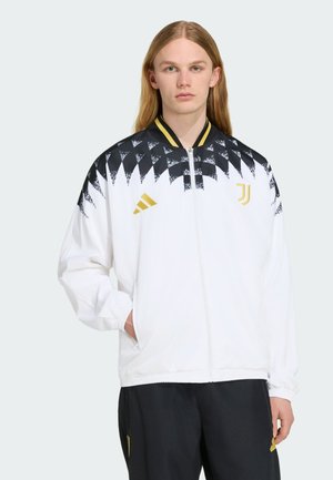 Jeune personne aux cheveux longs portant une veste de sport blanche et noire avec des logos dorés et un pantalon noir, debout devant un fond uni.