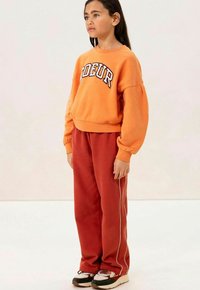 Een oranje sweatshirt met de tekst "CŒUR", gecombineerd met rode trainingsbroeken met witte zijstrepen en zwarte sneakers met een ontwerpdetail.