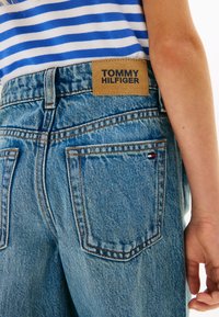 Ανοιχτό μπλε τζιν με τραχιά υφή, διακοσμημένο με καφέ δερμάτινη ετικέτα που φέρει το όνομα "TOMMY HILFIGER" και ραφές σε κόκκινο, λευκό και μπλε χρώμα.