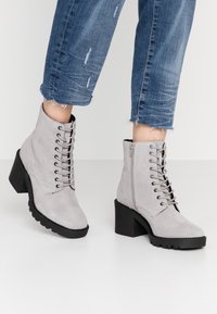 Botas de tobillo de ante gris con cordones en la parte delantera, cremallera lateral, tacón cuadrado y suela de goma texturizada. Llevadas con jeans azules recortados.