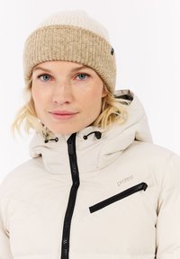 Beige Steppjacke mit schwarzem Reißverschluss und Brusttasche, kombiniert mit einer zweifarbigen Strickmütze in Beige und Creme. Weiche Textur, lässiges Design.