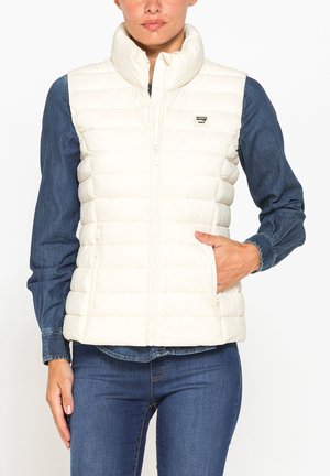 Donna che indossa un gilet smanicato bianco trapuntato sopra una camicia di jeans e jeans, con la mano nella tasca del gilet.