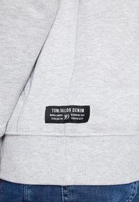Sweat-shirt gris clair avec une étiquette noire rectangulaire Tom Tailor Denim près de l'ourlet inférieur, associé à un jean bleu.