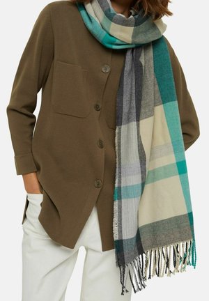 Femme portant un cardigan boutonné vert olive, un pantalon blanc et une grande écharpe à carreaux dans les tons bleu sarcelle, crème, gris et noir avec des franges aux extrémités.