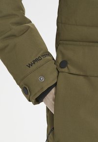 Olivgrön jacka med ett kviltat mönster, med svart muddar och tryckknappar, samt "W-PRO 10000" broderat på ärmen.