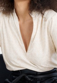 Světle béžová blůza s výstřihem do V, zdobená jemnými bílými vzory listů; materiál je hladký a lehký. Doplněna černým páskem.