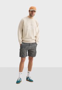 Sweatshirt beige avec texte imprimé, short cargo à motifs gris, chaussettes blanches avec accents colorés et baskets teal. Une casquette orange complète le look.