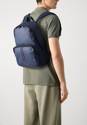 Zaino blu navy con un esterno in nylon liscio, cerniera a contrasto e un sottile dettaglio a strisce. Le spalline ergonomiche migliorano il comfort.