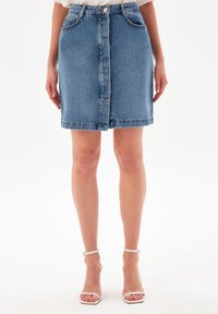 Kvinna som bär en blå jeans minikjol med framknappdetalj och vita öppna hälskor med ankelsnöre, stående mot en enfärgad vit bakgrund.