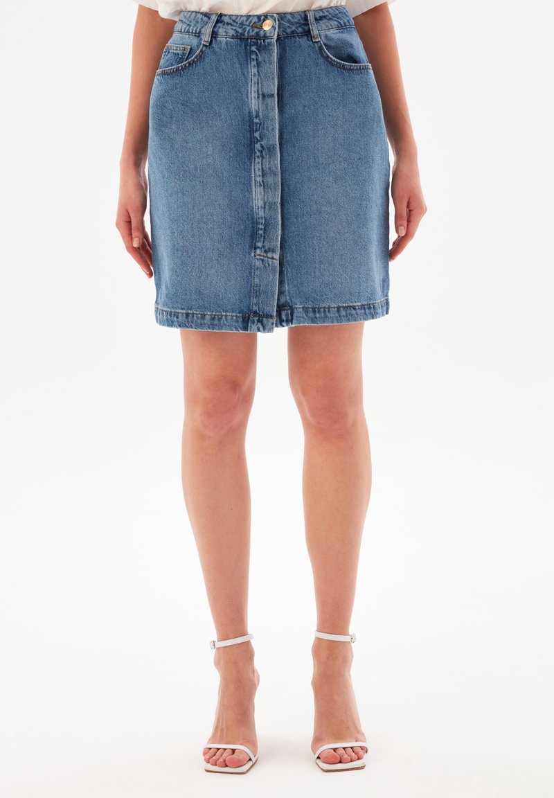 Kvinna som bär en blå jeans minikjol med framknappdetalj och vita öppna hälskor med ankelsnöre, stående mot en enfärgad vit bakgrund.