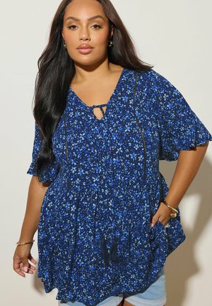 Pitkähiuksinen nainen, jolla on sininen kukallinen löysä pusero, jossa on tupsunarut, farkkushortsit, kultaiset rannekorut ja renkaankorvakorut, seisoo vaaleaa seinää vasten.