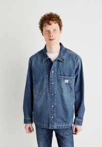 Camicia in denim blu scuro, con chiusura a bottoni, due tasche sul petto e etichetta con logo. Tessuto strutturato e vestibilità rilassata.