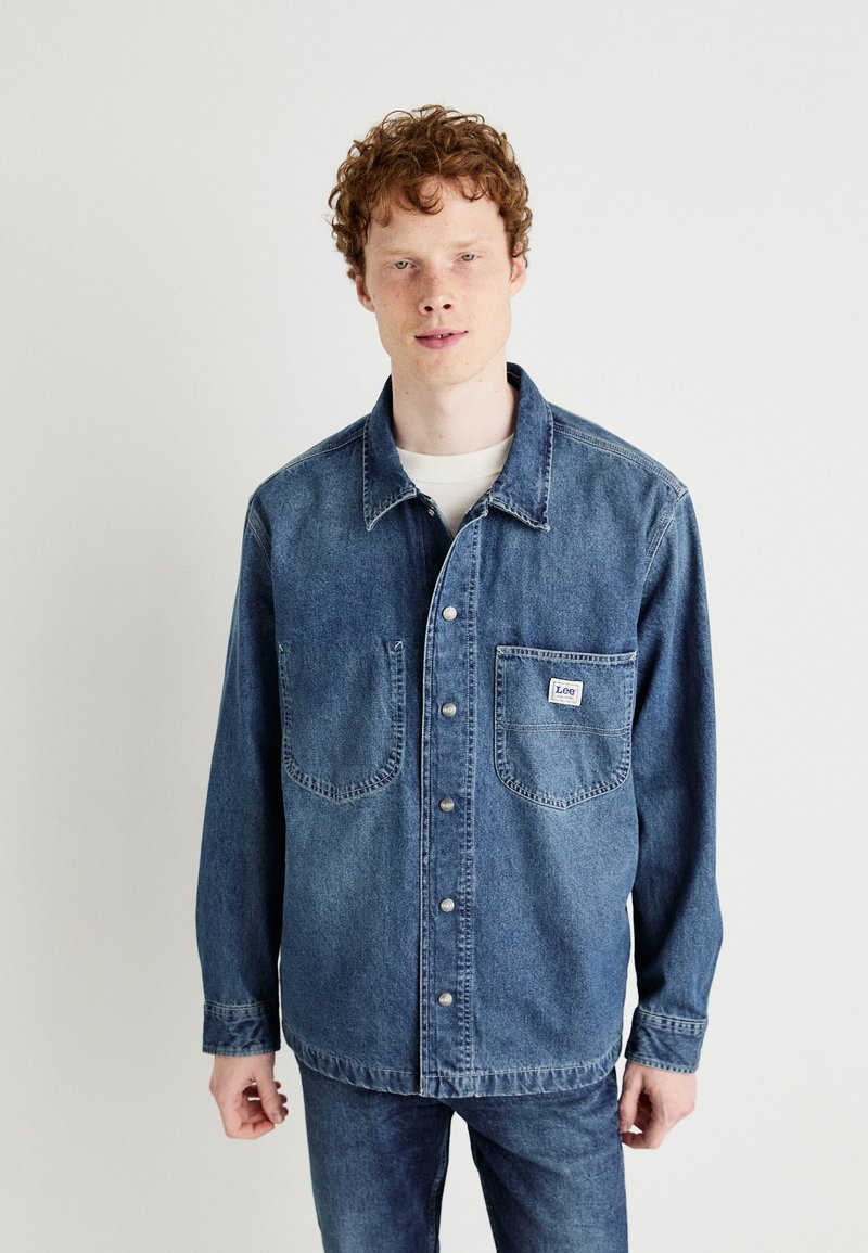 Camicia in denim blu scuro, con chiusura a bottoni, due tasche sul petto e etichetta con logo. Tessuto strutturato e vestibilità rilassata.