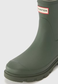 Botte de pluie en caoutchouc vert avec une pointe arrondie, semelle texturée et une étiquette de marque rouge et blanche sur la tige. Fini mat lisse.