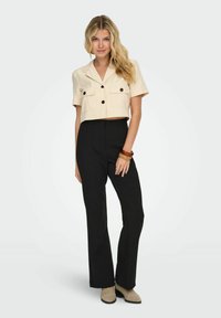 Chemise courte beige à manches courtes avec deux poches poitrine et des boutons noirs, associée à un pantalon noir taille haute et des bottes beiges.