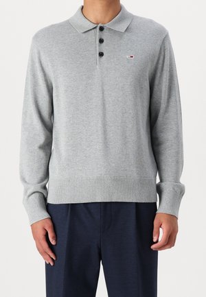 Muž nosící světle šedý polo svetr s dlouhým rukávem, černými knoflíky a malým logem na levé straně hrudi, v kombinaci s tmavě modrými kalhotami.