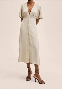 Robe midi en lin beige avec manches courtes bouffantes, un décolleté en V profond, une fermeture boutonnée, et une coupe décontractée. Associée à des talons hauts noirs à brides.