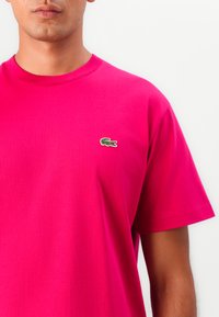 Fuchsia bomulds T-shirt med rund halsudskæring, korte ærmer og et lille grønt broderet krokodillelogo på venstre bryst.