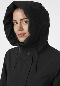 Černá kapuce s vysokým límcem, předním zipem a nastavitelnými šňůrkami pro bezpečné upevnění kolem obličeje. Hladká textura.