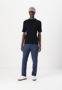 Marinblå stickad polo med korta ärmar, kombinerad med blå kostymbyxor och vita sneakers, kompletterad med en ljusgrå keps.