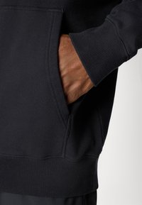 Sweatshirt preto com um bolso canguru, punhos e barra canelados. O tecido aparenta ser macio e texturizado. Mão parcialmente visível no bolso.