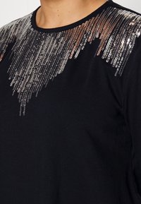 Svart tröja med metalliska paljetter i silver och brons i gradienter över axelpartiet, med en rundad halsringning.