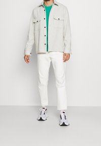 Chaqueta gris de botones con dos bolsillos en el pecho sobre una camiseta verde, combinada con pantalones blancos y zapatillas grises con detalles en negro.