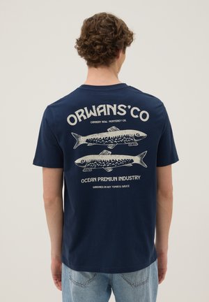 Person trägt ein dunkelblaues T-Shirt mit beigen Fischillustrationen und Text über Sardinen auf dem Rücken, kombiniert mit hellblauen Jeans.