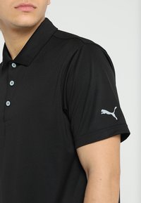 Mies, jolla on musta lyhythihainen poolopaita, jossa on kolme nappia ja valkoinen Puma-logo vasemmassa hihaosassa, yksinkertaista taustaa vasten.