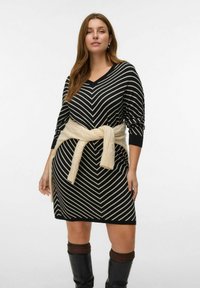 Robe en tricot noir et blanc à rayures avec un col en V et des manches longues, dotée d'une ceinture en tricot beige nouée à la taille. Bottes noires montantes.