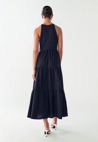 Žena s vlasmi v bunke, oblečená v bezrukávových námorníckych tiered maxi šatách a bielych podpätkových mule topánkach, stojaca na pozadí jednoduchého bieleho pozadia.