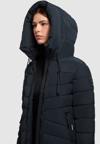 Sötétkék puffert jaket nagy kapucnival, steppelt designnal, elől cipzárral és húzózsinórokkal. Oldalán cipzáros zsebekkel és sima textúrával rendelkezik.