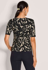 Vrouw met donker haar in een paardenstaart, gekleed in een zwart en beige abstract patroon kort mouwtop en zwarte aangesloten broek, gezien van achteren.