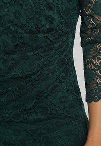 Robe en dentelle vert foncé avec motifs floraux et manches trois-quarts à bord festonné. Tissu texturé ajoutant de la profondeur.