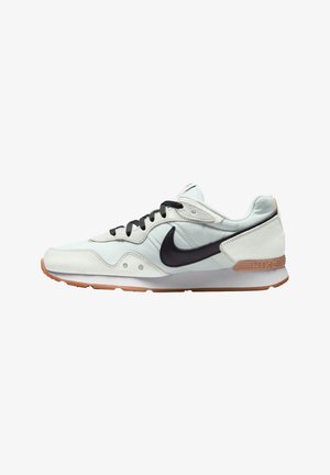 Biało-beżowy sneaker Nike z czarnym logo słynnego znaku, czarnymi sznurówkami i jasno brązową podeszwą, pokazany z boku na białym tle.
