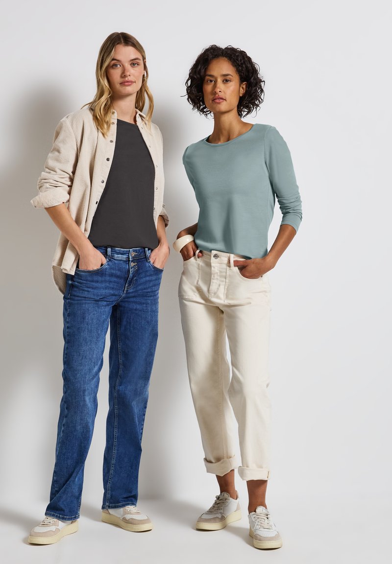 Dos mujeres están de pie una al lado de la otra. Una lleva una camisa gris y un cárdigan beige sobre unos jeans azules, mientras que la otra viste una camisa azul verdosa y pantalones blancos.
