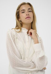Hvid knap-opp blouse med flæsedetaljer, teksturerede polkaprikker og plisseret front. Har lange, luftige ærmer med stribede manchetter.