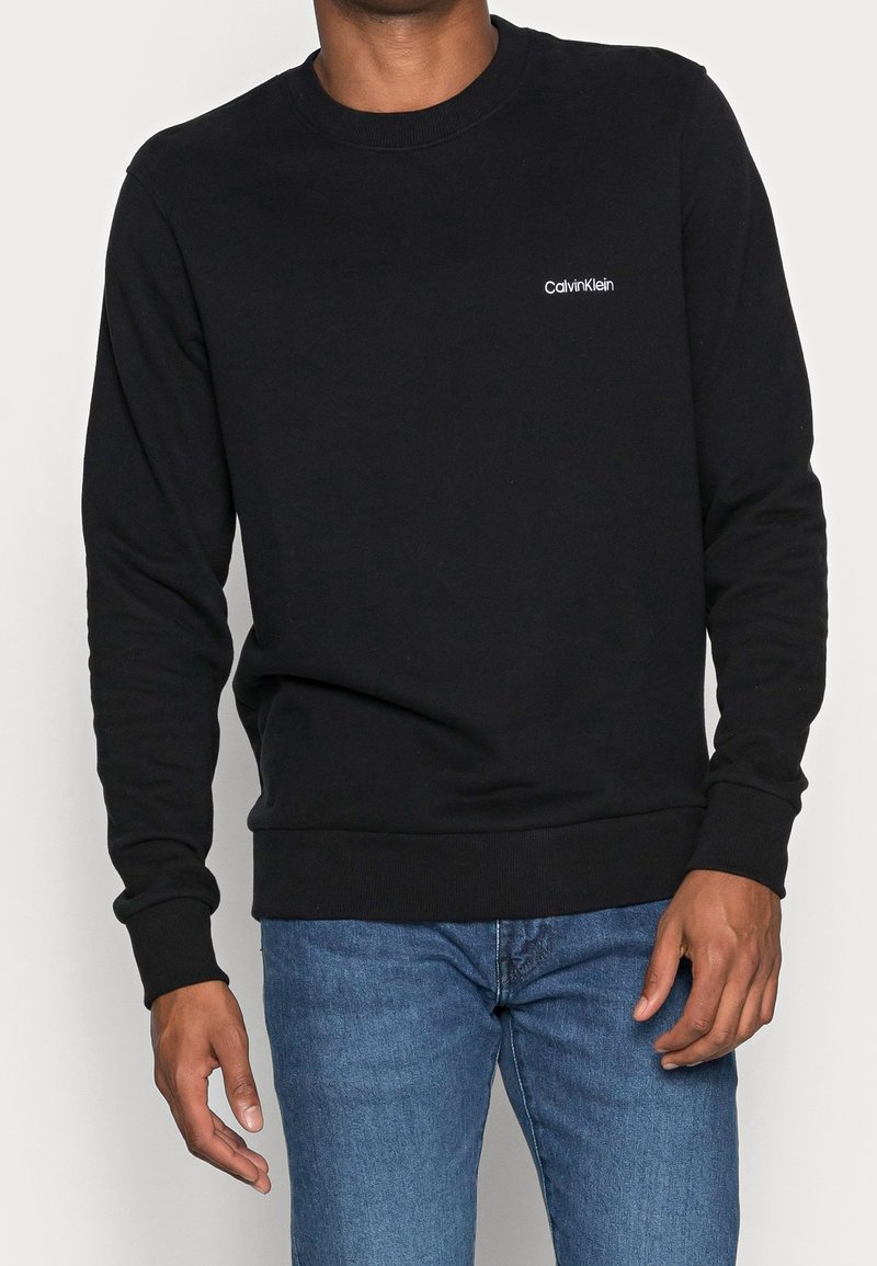Felpa nera in cotone con maniche lunghe, colletto a girocollo e polsini a costine. Presenta un piccolo logo bianco di Calvin Klein sul petto.