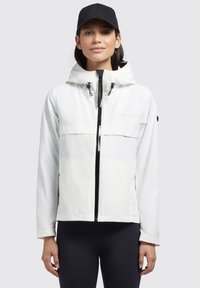 Veste à capuche blanche avec une fermeture éclair et des accents de poche noirs. Portée avec une casquette noire, mettant en avant un design élégant et fonctionnel.