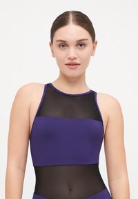 Haut de sport violet avec un panneau en maille noire texturée et des sections violettes unies, conçu avec un col haut et des bretelles fines.