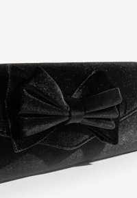 Melna teksturēta auduma rokassoma ar lielu satīna loku priekšpusē. Virsma ir viegli mirdzoša, taisnstūrveida formā.