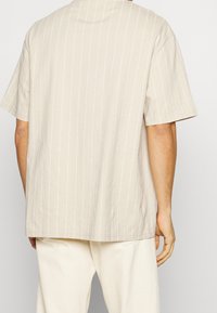 Krémové tričko s krátkým rukávem a bílými vertikálními pruhy, volný střih a kulatý výstřih. Měkká bavlněná tkanina s hladkou strukturou.