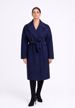 Frau trägt einen knie-, dunkelblauen Wollmantel mit Gürtel, schwarze Leggings und schwarze Stiefeletten mit Absatz, stehend vor weißem Hintergrund.