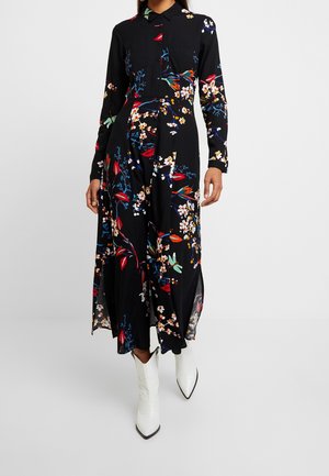 Woman wearing a long-sleeve black floral shirt dress with white pointed-toe boots, standing against a plain gray background.