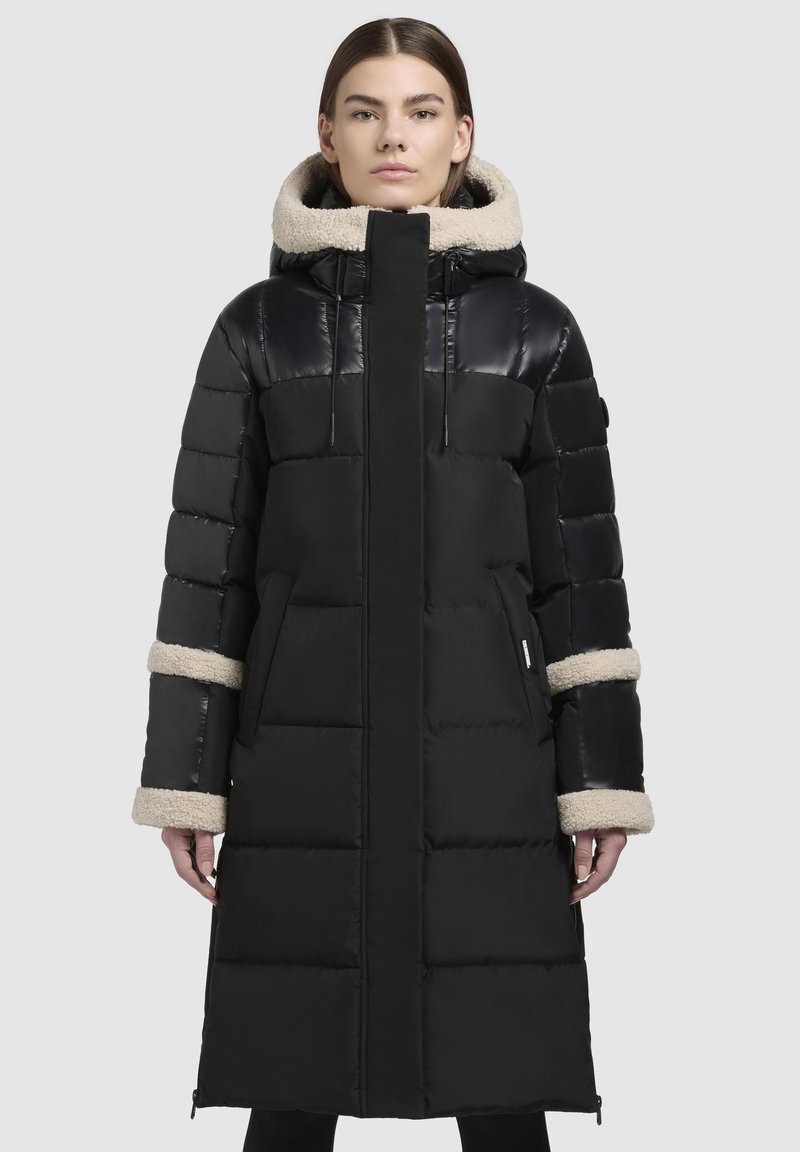 Schwarze Pufferjacke mit langem Design, glänzenden oberen Paneelen, gerippter Textur und einem flauschigen beigen Kapuze sowie Bündchen. Verfügt über Seitentaschen und Reißverschlüsse.