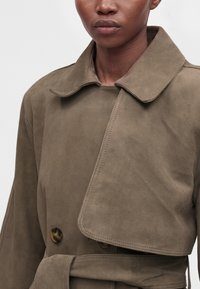 Gros plan d'une personne portant un trench-coat en daim taupe avec un grand col et des boutons en écaille de tortue, ceinture nouée à la taille.