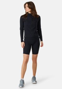 Giacca sportiva nera con zip frontale, abbinata a pantaloni da ciclismo neri. Indossata con sneaker grigie, caratterizzate da un design in tessuto testurizzato.