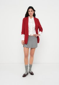 Κόκκινο σακάκι ραμμένο στο μέτρο, λευκό πουκάμισο με κορδόνια, γκρι πλισέ φούστα, γκρι κάλτσες, σκούρα μπορντό παπούτσια. Μινιμαλιστικός σχεδιασμός με στρωματοποιημένες υφές.